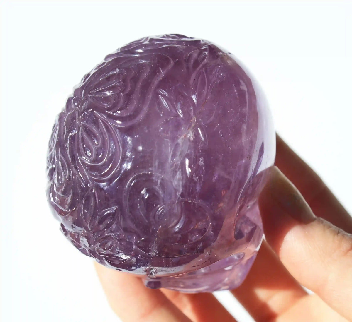 A hand holding a carved purple amethyst crystal skull against a bright white background.