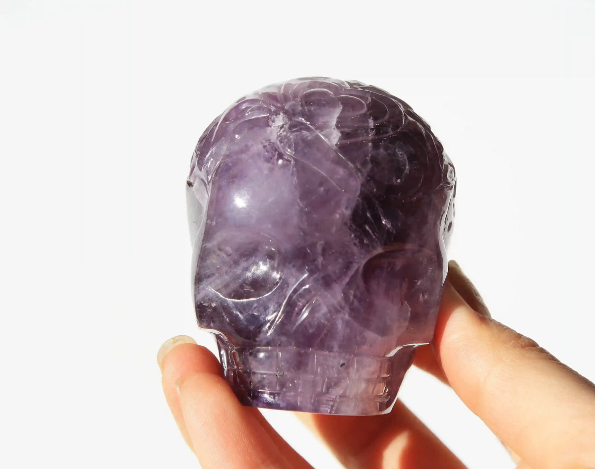 A hand holding a carved amethyst crystal skull against a bright white background.