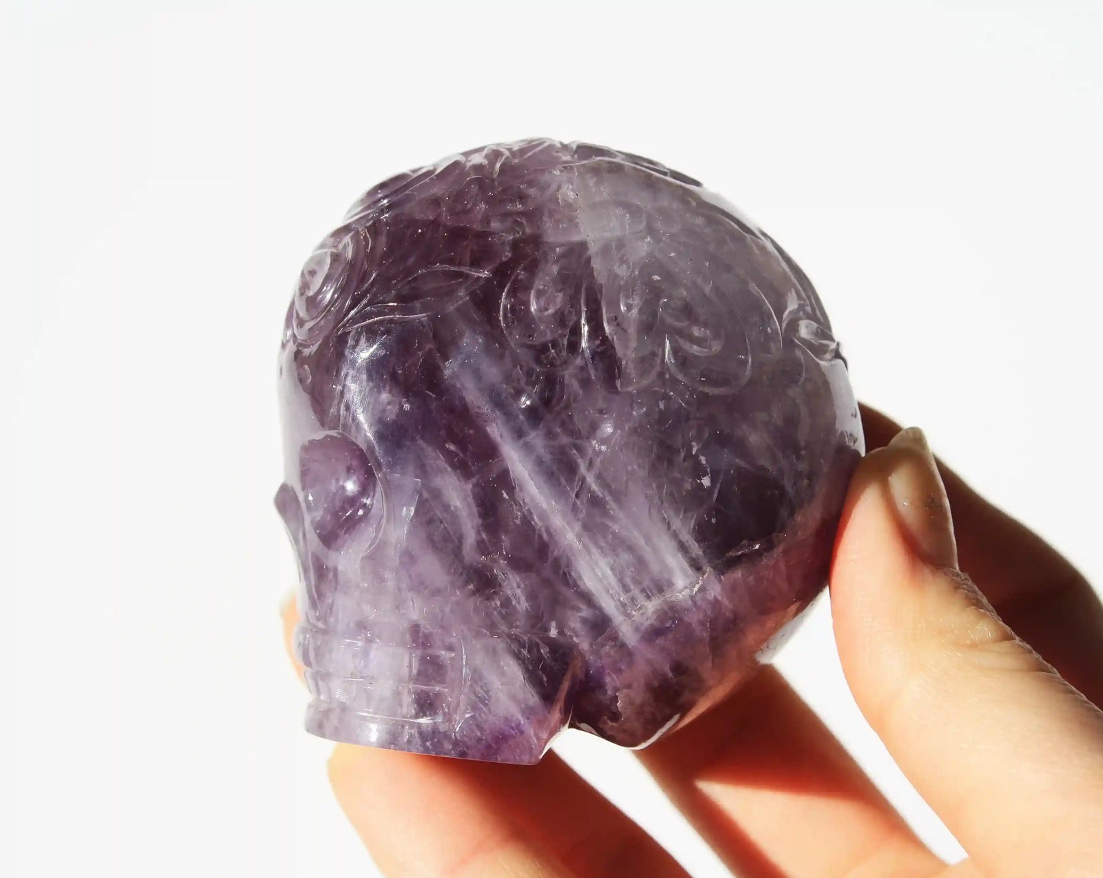 A hand holding a carved amethyst crystal skull against a bright white background.