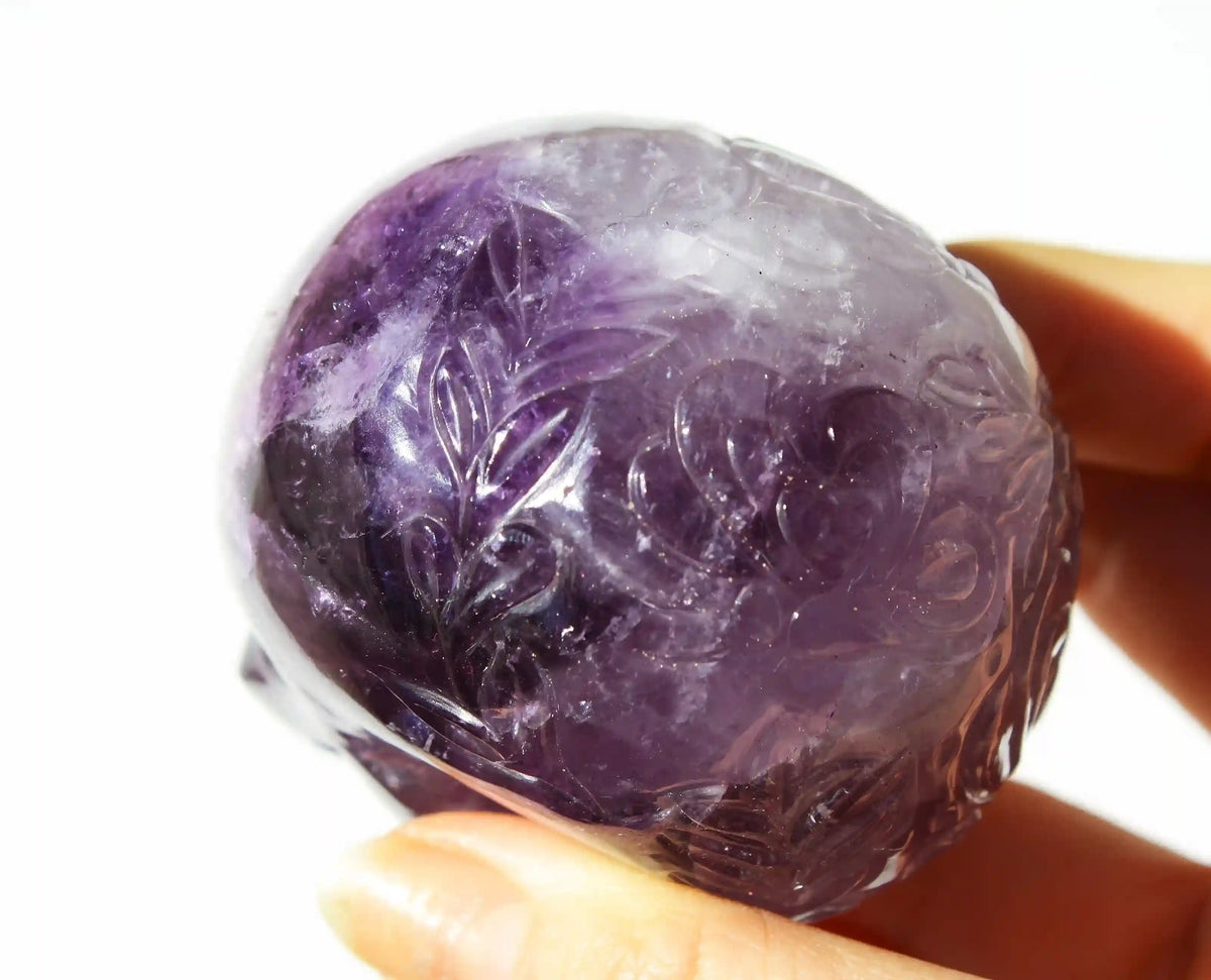 A hand holding a carved amethyst crystal skull against a bright white 